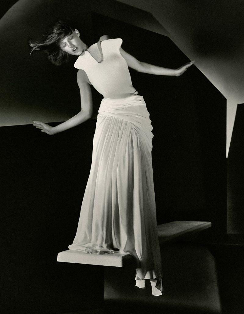 Model in a white sleeveless top and pleated long skirt posing dramatically on a raised architectural platform