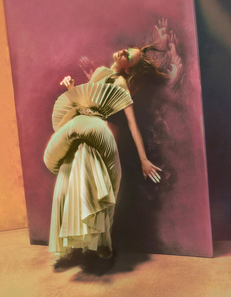 Model in architectural pleated dress leaning against a magenta wall with painted hands