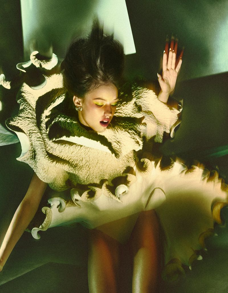 Model in a sculptural ruffled beige dress with dramatic movement captured against a muted greenish architectural background