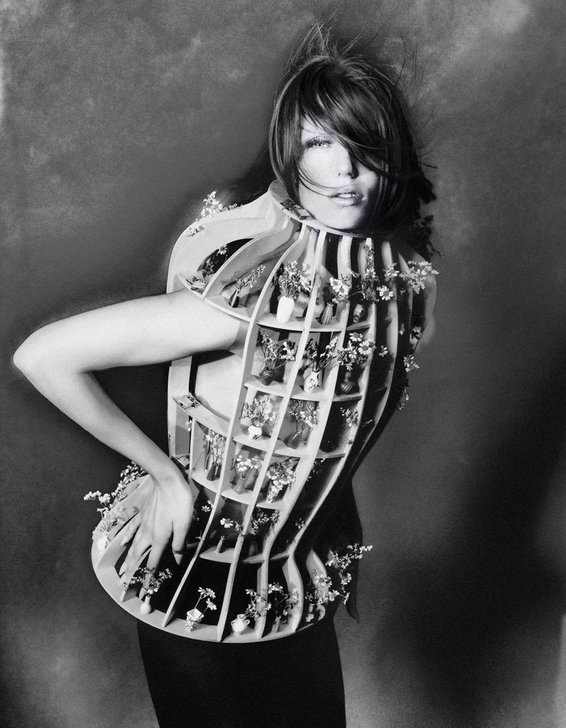Monochrome fashion editorial portrait of a female model wearing an architectural wooden cage garment filled with tiny flowers
