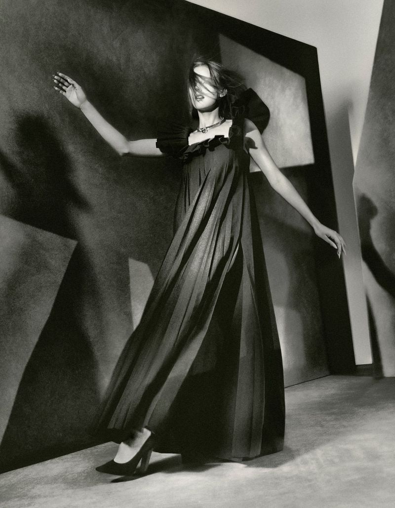 Model wearing a dark pleated maxi dress and high heels posing dynamically against an abstract sculptural backdrop.