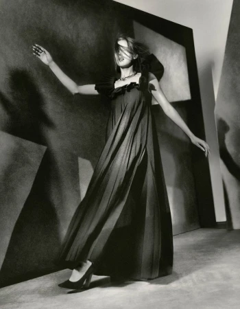 Model wearing a dark pleated maxi dress and high heels posing dynamically against an abstract sculptural backdrop.