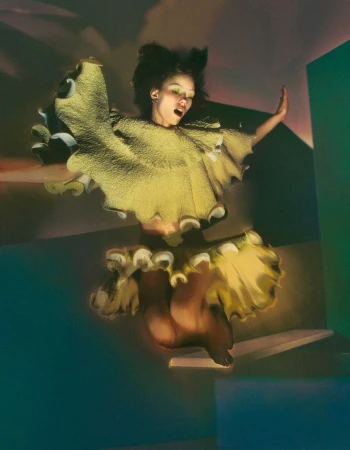 Model in a sculptural yellow ruffled top jumping mid-air in a stylized set by Elizaveta Porodina for T Magazine Germany