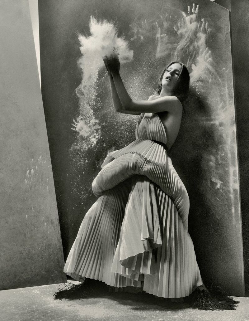 Model in a sculptural pleated dress standing against a dark textured backdrop with an explosion of white powder