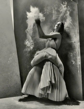 Model in a sculptural pleated dress standing against a dark textured backdrop with an explosion of white powder