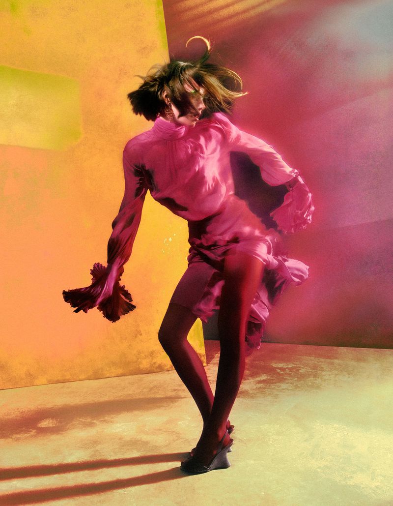 Model in dynamic pose wearing a bright pink blouse and skirt against a vibrant yellow and pink painted backdrop.