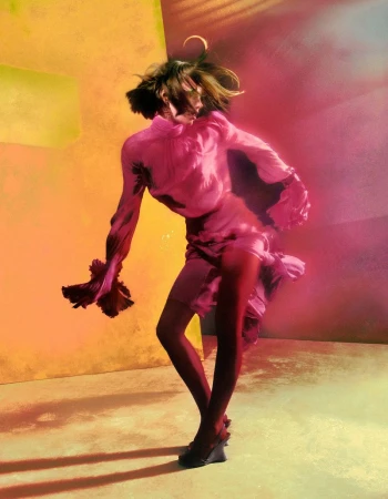 Model in dynamic pose wearing a bright pink blouse and skirt against a vibrant yellow and pink painted backdrop.