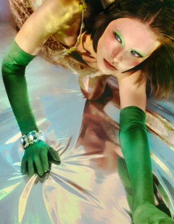 Model in green opera gloves and metallic dress leaning over reflective surface with bright green eyeshadow.