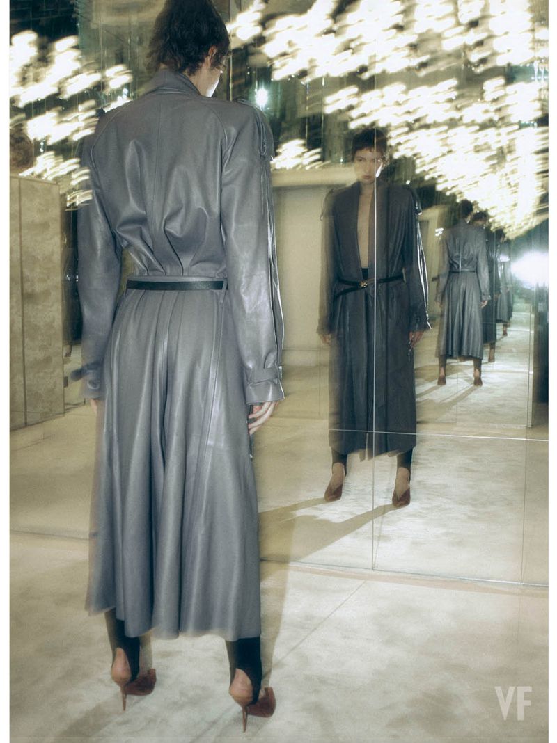 Model wearing a grey belted trench coat standing in front of a large mirror reflecting multiple figures.