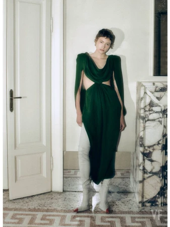 Model with short hair wearing a dark green cutout dress and white boots standing in a hallway