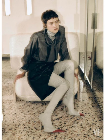 Model with short dark hair wearing a sheer grey blouse and white thigh-high boots seated in an armchair