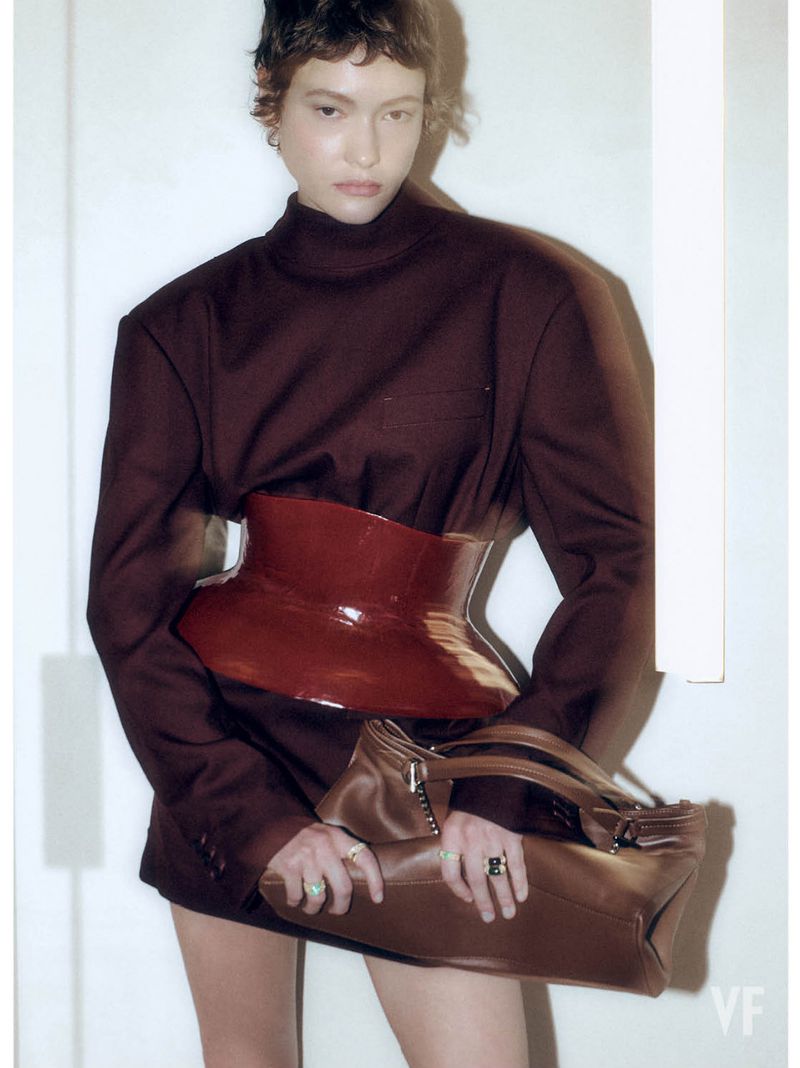 Model in structured dark top wearing a glossy red waist belt and holding a brown leather handbag against white background