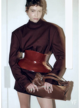 Model in structured dark top wearing a glossy red waist belt and holding a brown leather handbag against white background