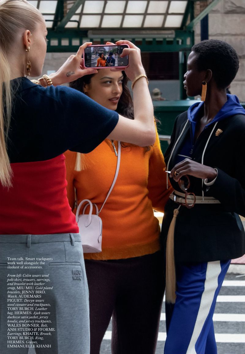 Three women in casual street fashion are gathered together; one woman is using a smartphone to capture a photo outdoors.