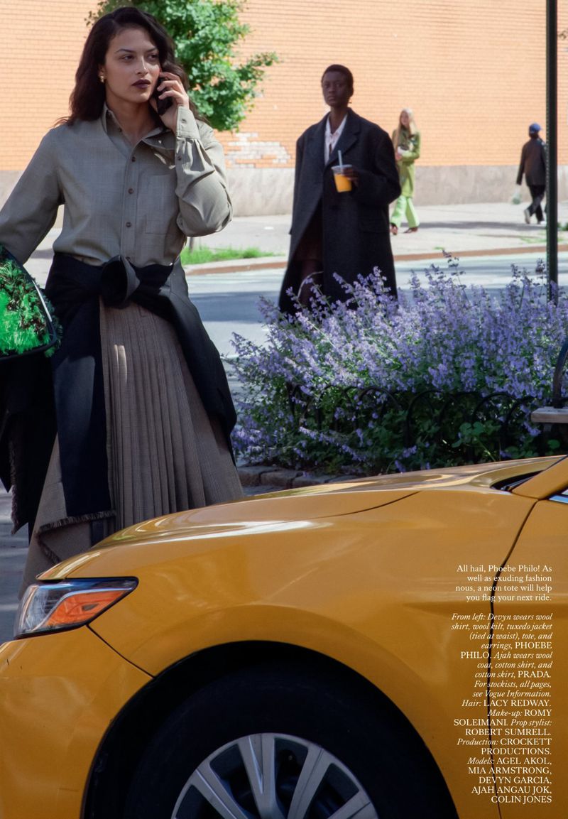 Woman talking on phone stands by yellow taxi on a city street. She wears olive shirt, pleated skirt, with buildings, trees and other people blurred.