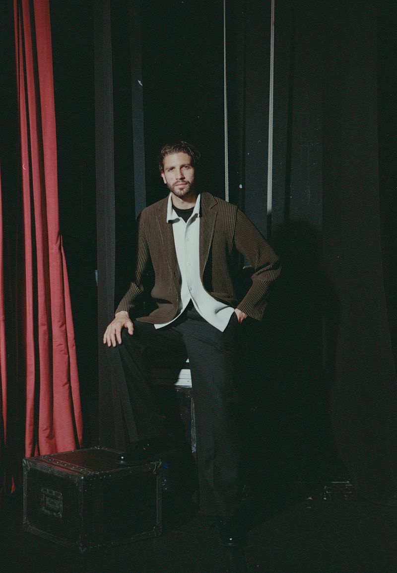 Man wearing a brown textured jacket and white collared shirt standing against a dark background with red curtains