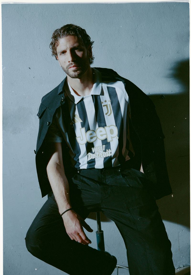 Manuel Locatelli wearing a black and white striped jersey and dark trousers seated against a textured grey wall