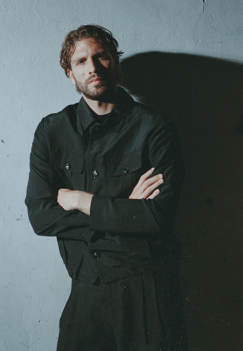Man with beard and dark hair wearing a black jacket with crossed arms posing against a grey wall with a strong shadow cast behind him