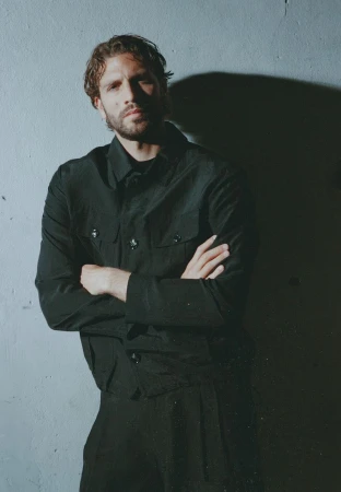 Man with beard and dark hair wearing a black jacket with crossed arms posing against a grey wall with a strong shadow cast behind him