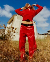 Model with curly red hair wearing a bright red top and matching red leather trousers outdoors in a dry field.