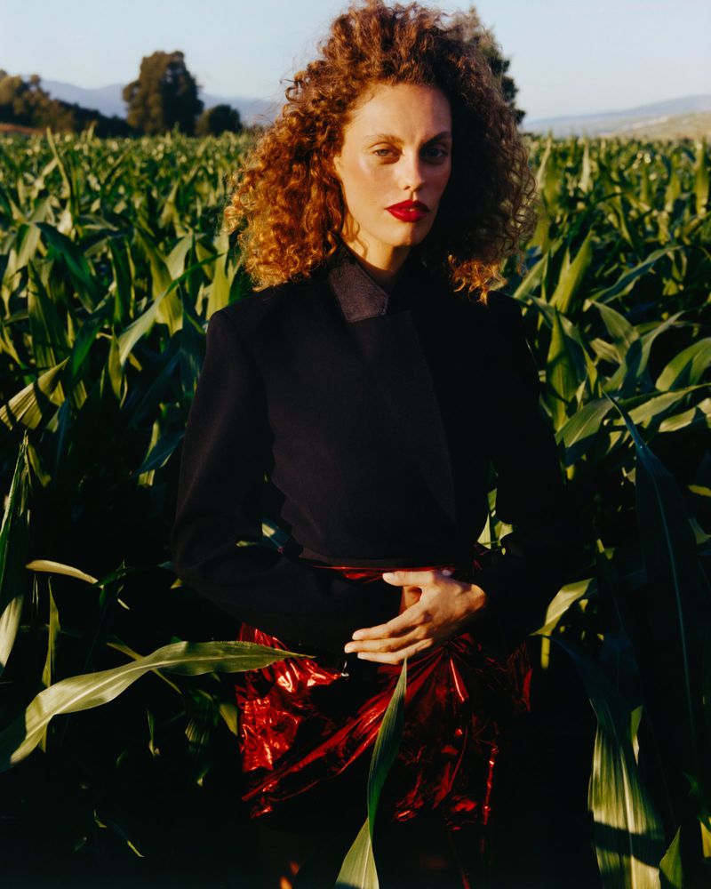 Model with curly red hair wearing a dark jacket and red metallic skirt standing in a tall green cornfield outdoors.