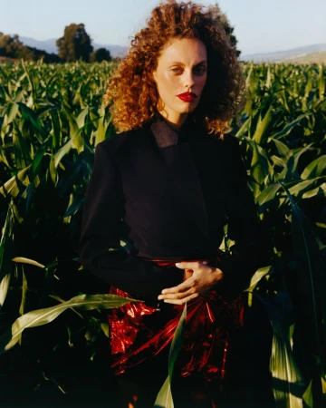 Model with curly red hair wearing a dark jacket and red metallic skirt standing in a tall green cornfield outdoors.