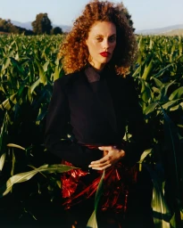 Model with curly red hair wearing a dark jacket and red metallic skirt standing in a tall green cornfield outdoors.