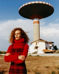 Model with curly red hair wearing a red turtleneck under a plaid dress in front of a large water tower.