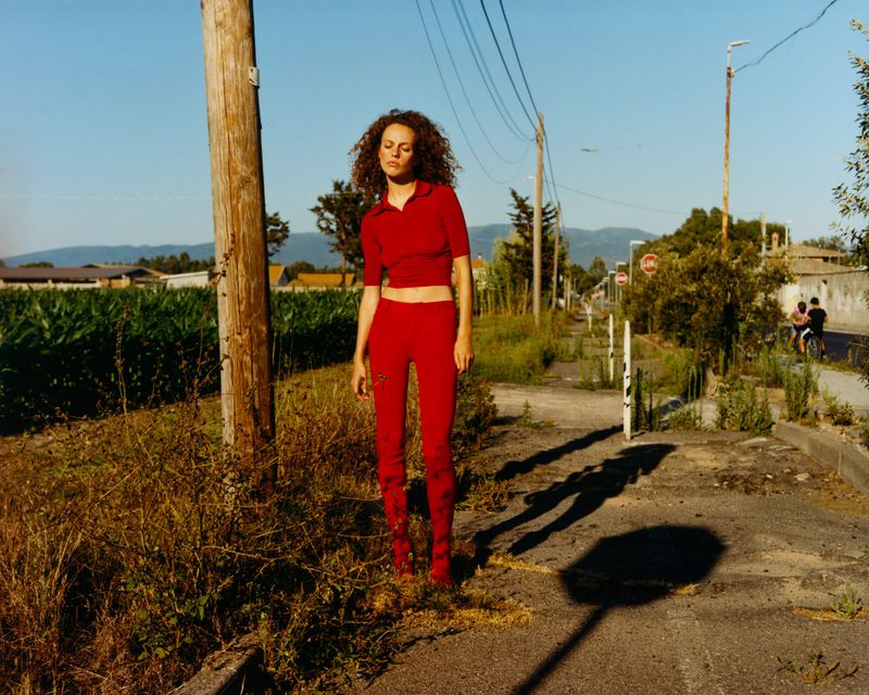 Model with curly hair wearing a red polo crop top and matching trousers standing on a rural roadside under daylight.