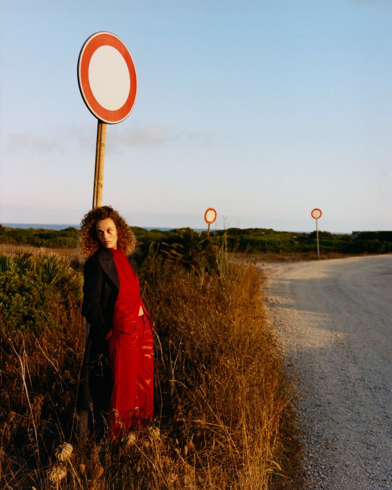 Model Gemma Janes in red trousers and black coat leaning by a road sign under a clear sky for iO Donna editorial Rouge Noir