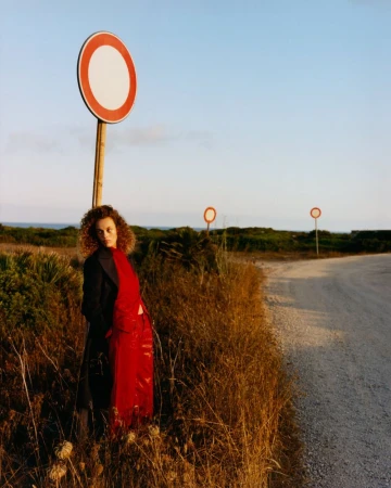 Model Gemma Janes in red trousers and black coat leaning by a road sign under a clear sky for iO Donna editorial Rouge Noir