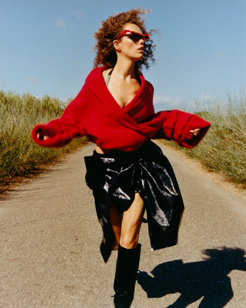 Model with curly hair wearing a red sweater and black skirt running along an outdoor road under a clear sky.