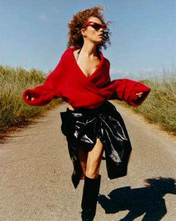 Model with curly hair wearing a red sweater and black skirt running along an outdoor road under a clear sky.