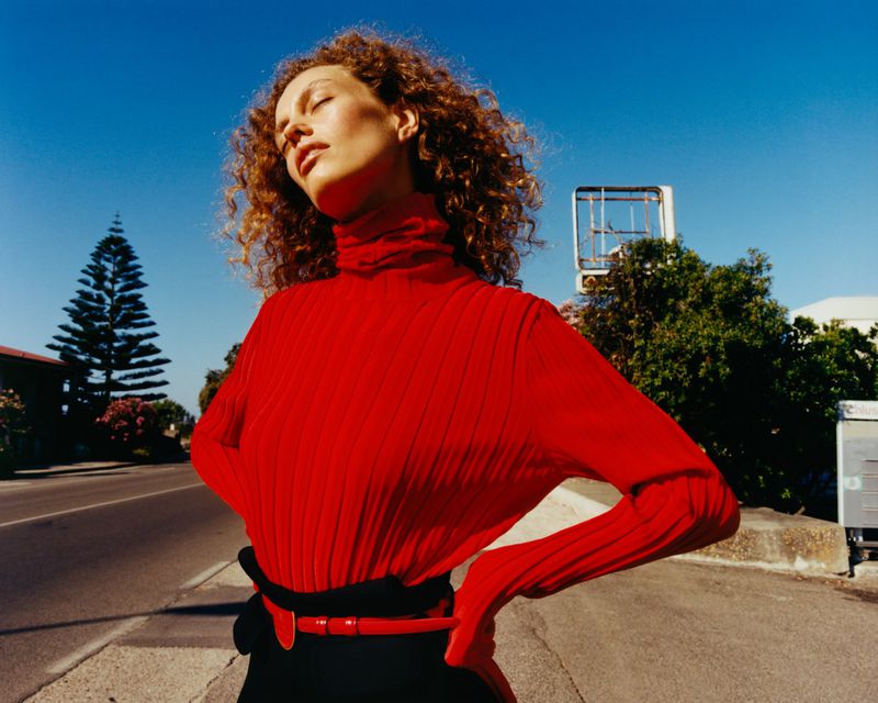 Model with curly hair wearing a red turtleneck sweater posing outdoors on a street under a clear blue sky