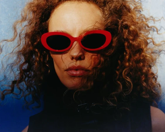 Close-up portrait of a woman with curly brown hair wearing bright red oval sunglasses against a blue background