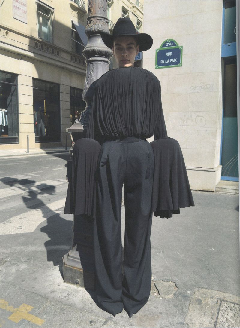 Model in black hat and pleated black outfit standing on a city street beside a lamp post.