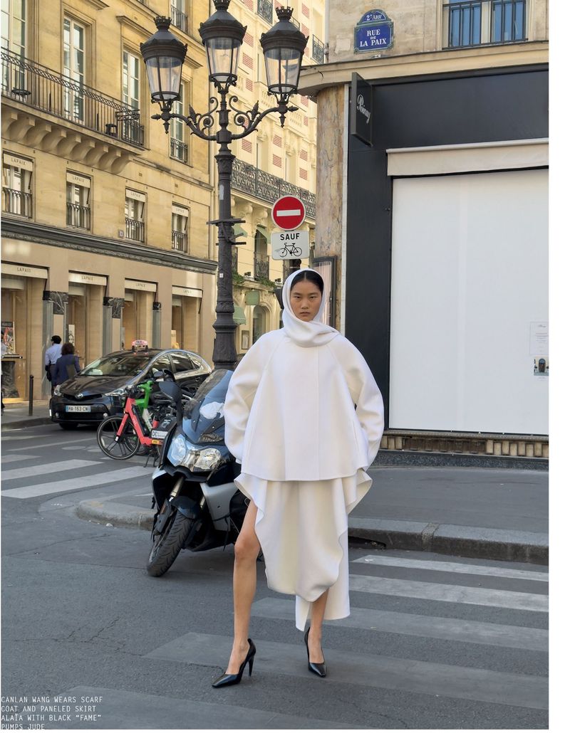 Model in white hooded coat and skirt standing on a Parisian street with a motorcycle and lamppost
