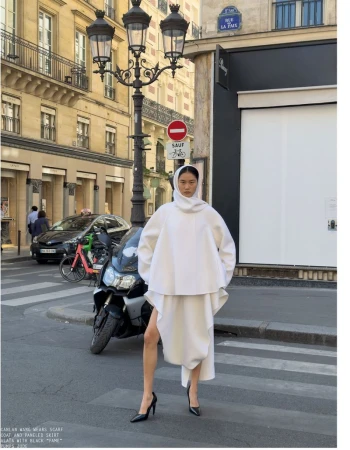 Model in white hooded coat and skirt standing on a Parisian street with a motorcycle and lamppost