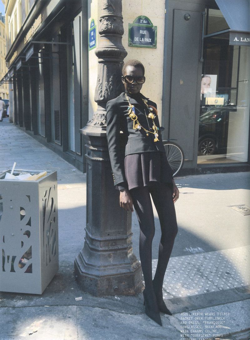 Model in black sunglasses leaning against a lamppost on a city street wearing a fitted black jacket and dark tights