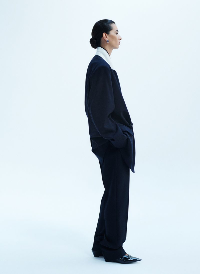 Model standing in profile wearing an oversized dark suit jacket and trousers against a seamless white background.