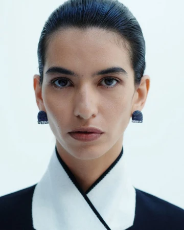 Close-up portrait of a model with dark slicked-back hair wearing dark blue crystal earrings against a white background.