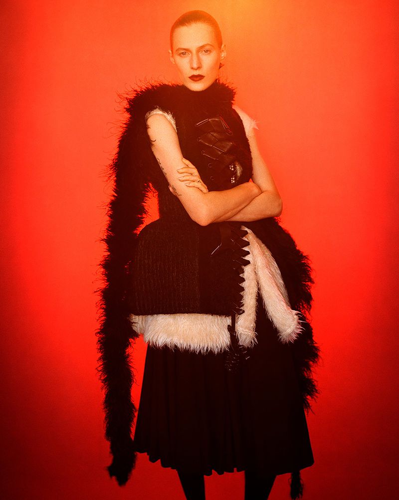 Model Julia Nobis wearing black feather-trimmed garments with arms crossed against vibrant orange gradient backdrop