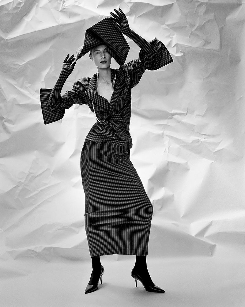 Model Julia Nobis in pinstripe skirt suit and sculptural hat against a crinkled white backdrop for W Magazine