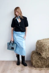 Model standing against a white wall wearing a black zip jacket, blue skirt, and black boots while holding a patterned tote bag