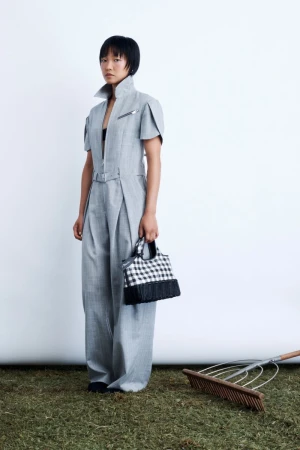Model standing on grass in a grey jumpsuit holding a black and white checkered handbag with a garden rake nearby against a white backdrop