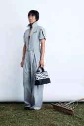 Model standing on grass in a grey jumpsuit holding a black and white checkered handbag with a garden rake nearby against a white backdrop