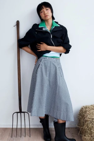 Model posing against white wall with pitchfork wearing black jacket and gingham skirt