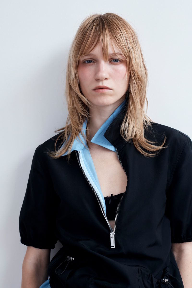 Model with blonde layered hair wearing a black zip top with a light blue collar against a white background