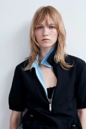 Model with blonde layered hair wearing a black zip top with a light blue collar against a white background