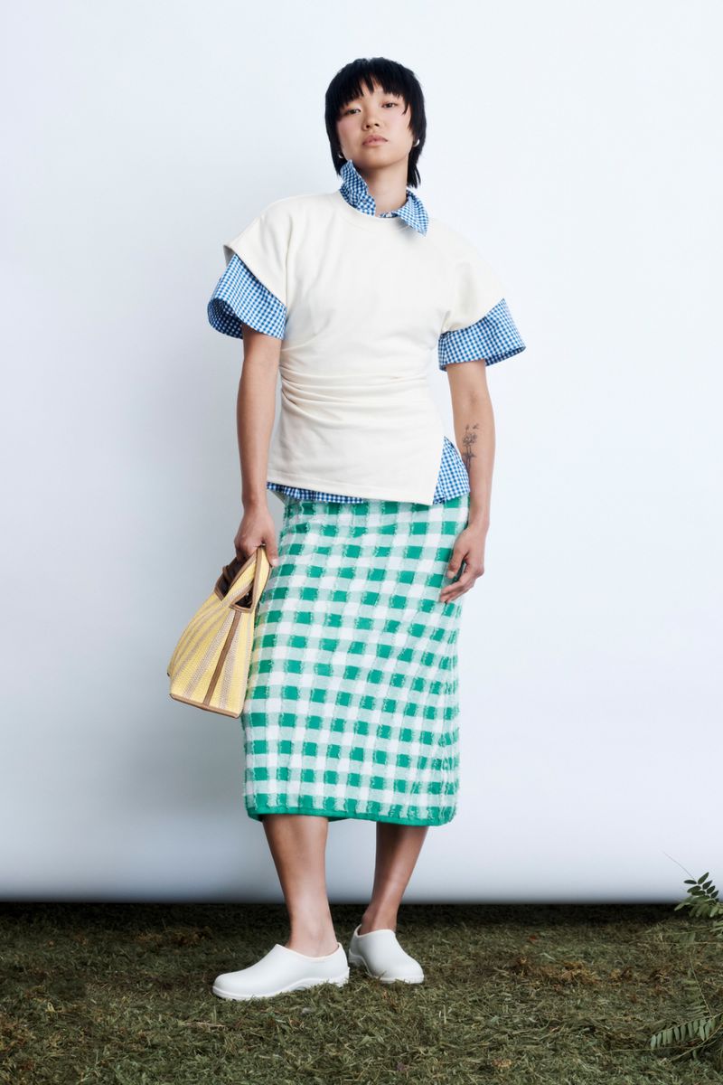 Model standing against a white backdrop wearing a white top over a blue gingham shirt, green gingham skirt, and holding a yellow handbag